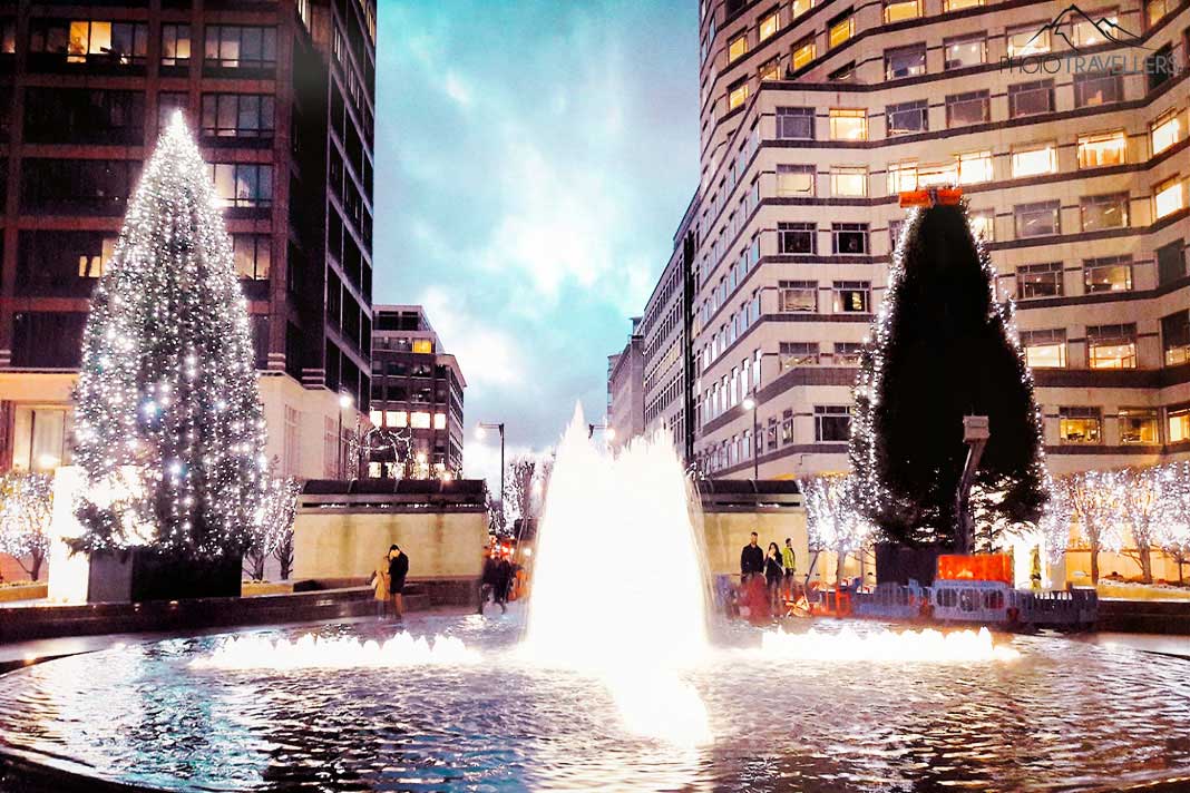 Beleuchtete Wasserfontäne und Christbäume vor den Hochhäusern in Canary Wharf