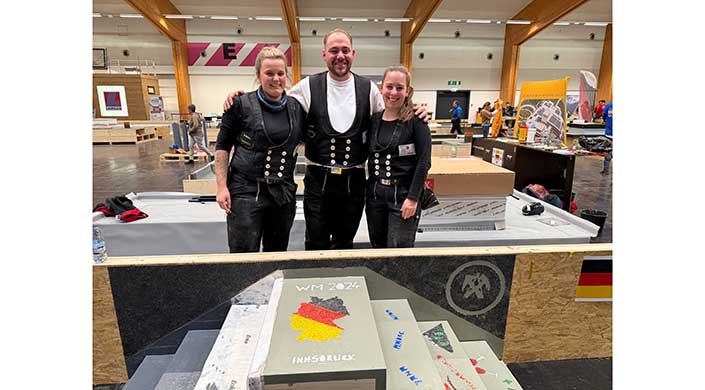 Drei Menschen in traditioneller Zimmermannskleidung stehen zusammen und lächeln. Im Vordergrund liegt ein Buch mit WM 2024 und einer Deutschlandkarte darauf.