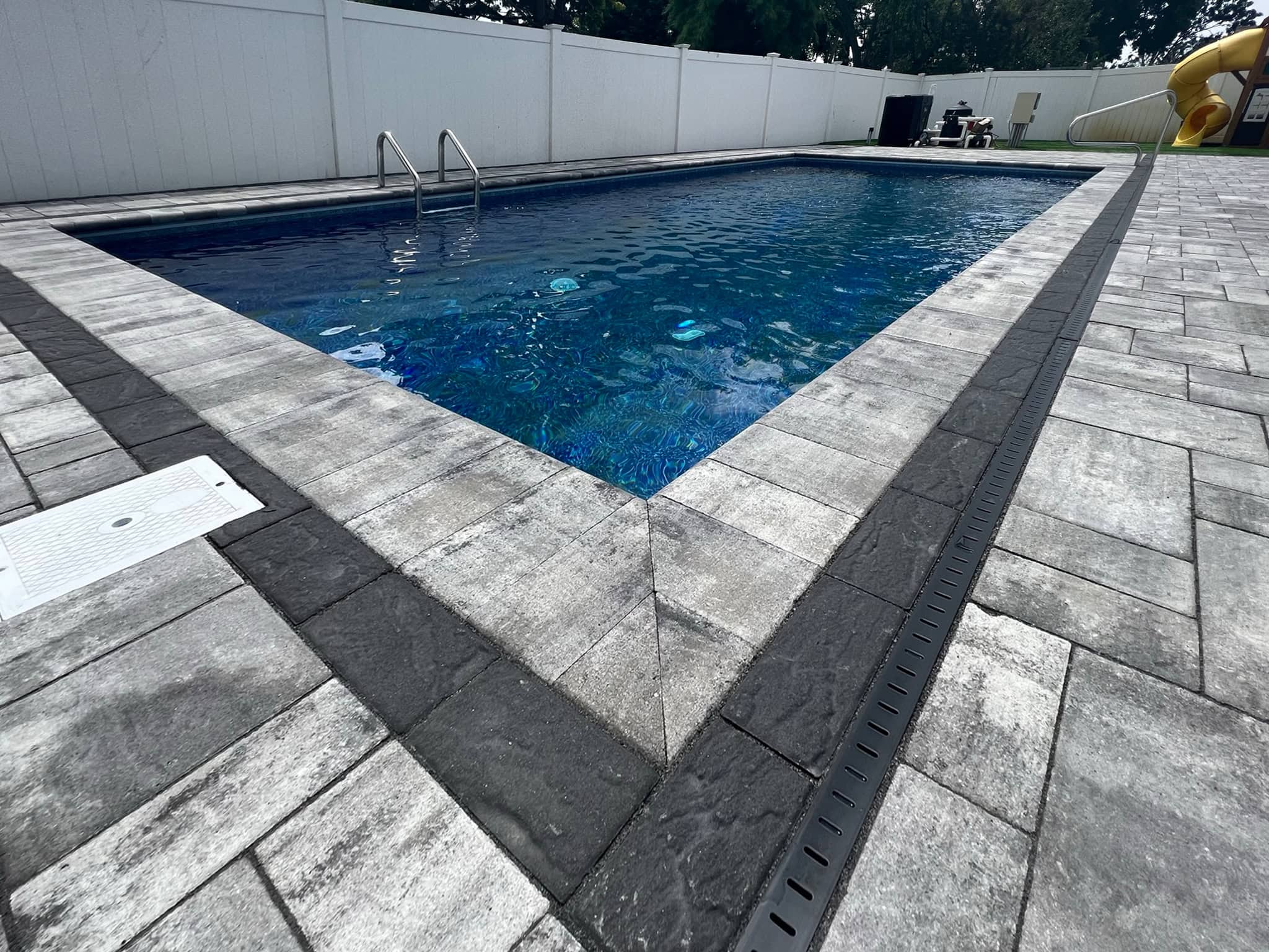 Rectangular outdoor swimming pool with metal ladder, surrounded by gray stone tile and a white privacy fence.