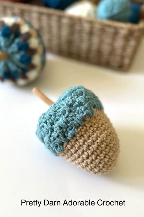 Beige and blue crocheted acorn on white surface; blurred basket of crochet items in background. Text: Pretty Darn Adorable Crochet. Free crochet pattern