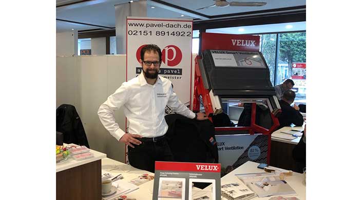 Ein Mann mit Bart steht an einem Messestand mit Werbematerialien, darunter Banner und Prospekte, die für Dachdeckerarbeiten und VELUX-Produkte werben.