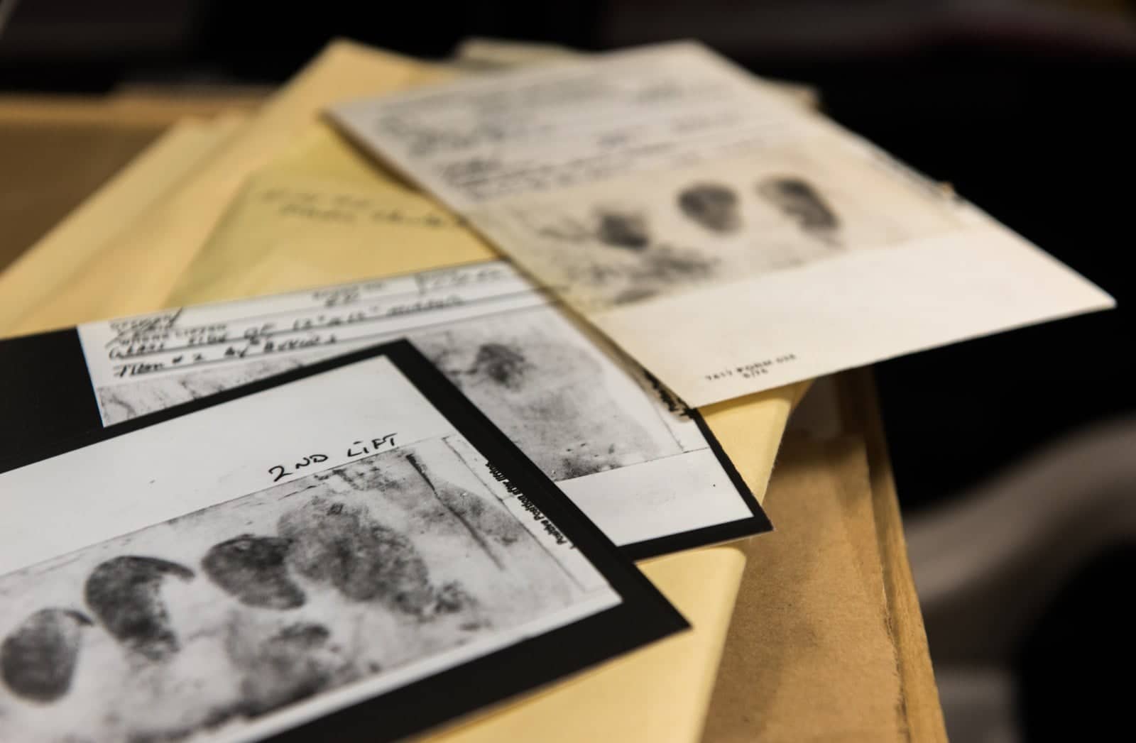 Several old fingerprint lift cards with dark smudged prints, spread on a table among evidence envelopes.