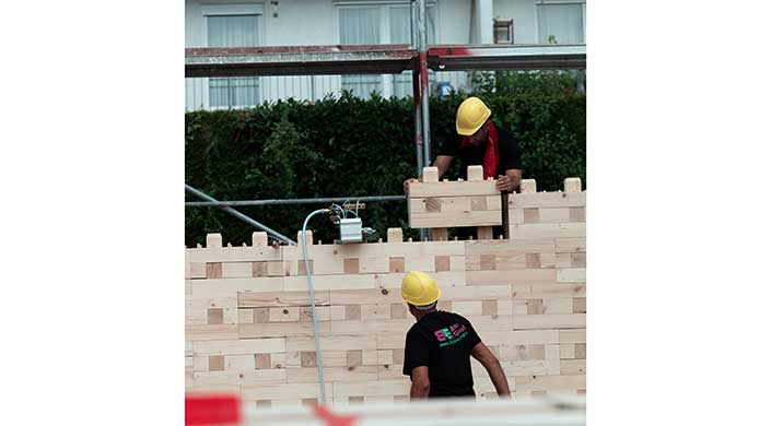 Zwei Bauarbeiter mit gelben Helmen arbeiten an einer Holzkonstruktion im Freien. Ein Haus oder Gerüst ist im Hintergrund sichtbar.