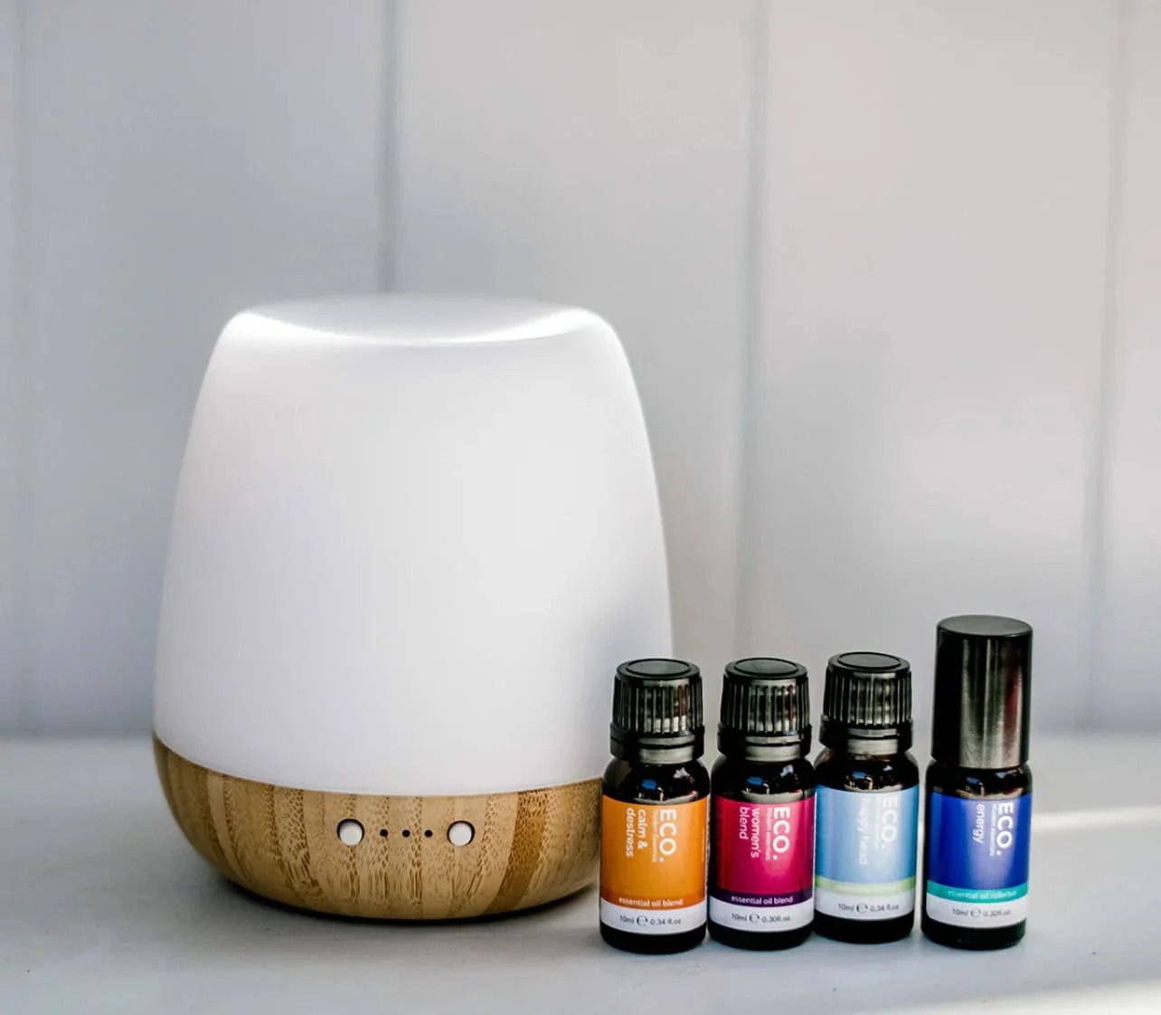 Revive & Thrive Aromatherapy Bundle 13 The Revive & Thrive Aromatherapy Bundle features a white and bamboo essential oil diffuser beside four essential oils with colorful labels, all arranged on a white surface against a light gray background—ideal for enhancing your aromatherapy experience.