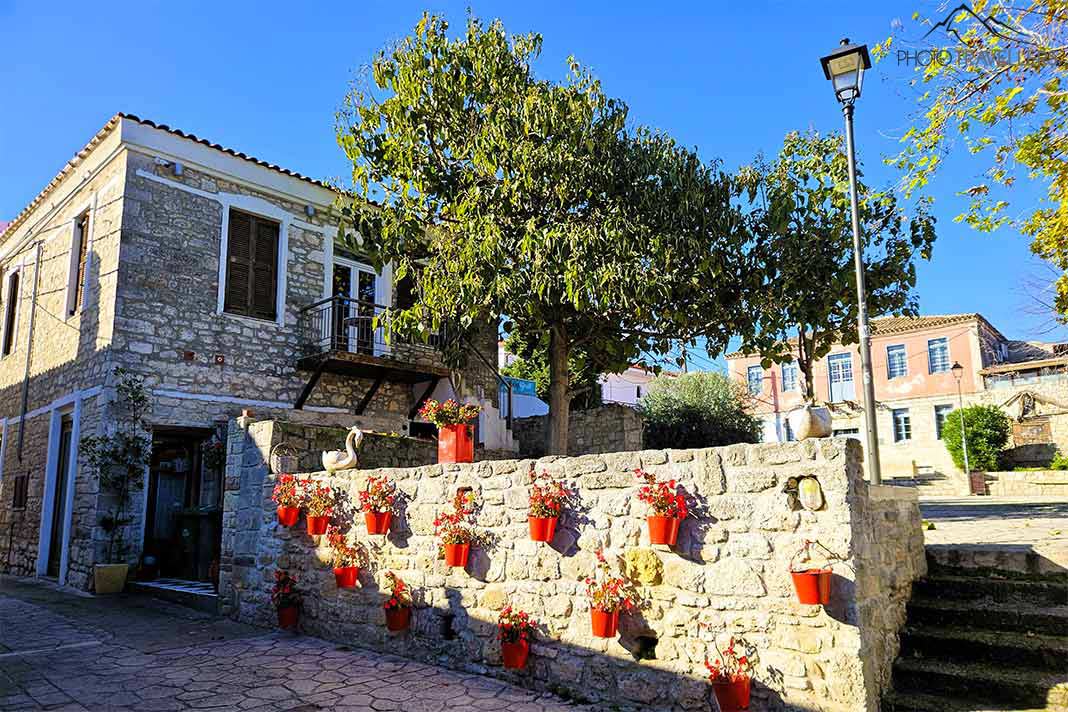 Rote Blumenkübel zieren eine Steinmauer, dahinter ein Baum und ein altes Steinhaus