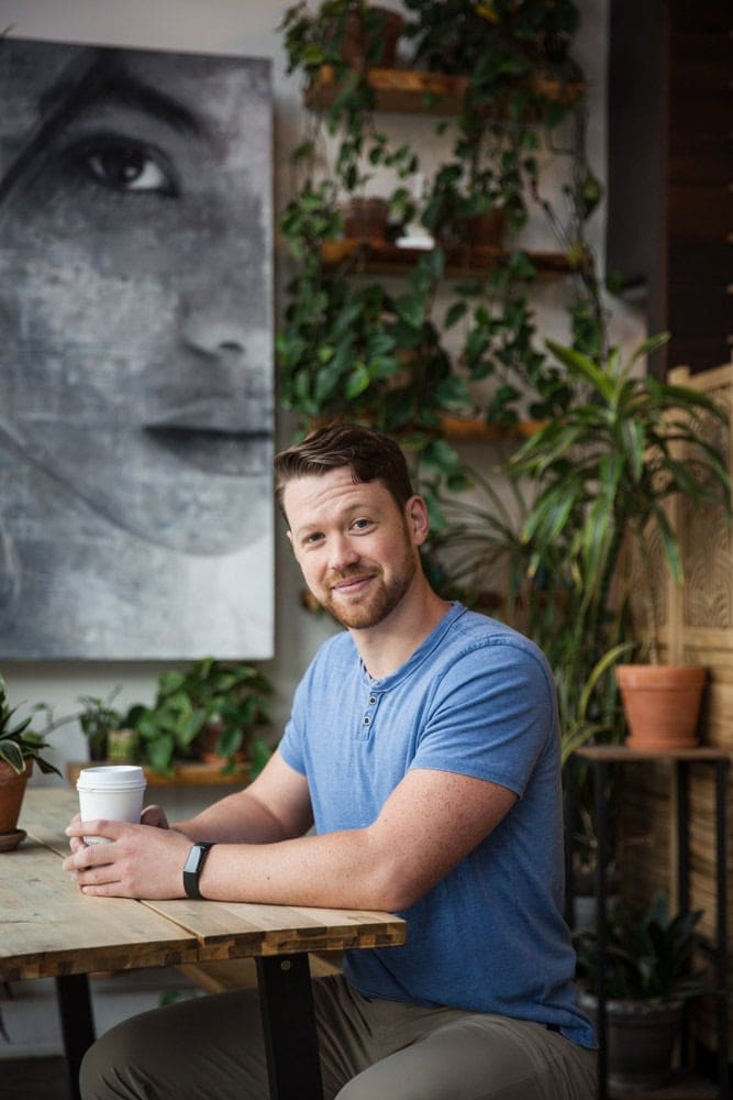 A man with short hair and a beard sits at a wooden table holding a coffee cup, surrounded by potted plants, with a large grayscale portrait in the background.