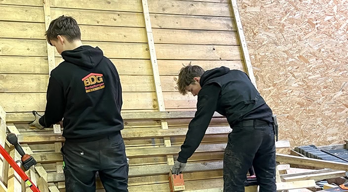 Junger Mann arbeitet mit einer Holzlatte und einer Werkzeugkiste vor einer Holzplankenwand.