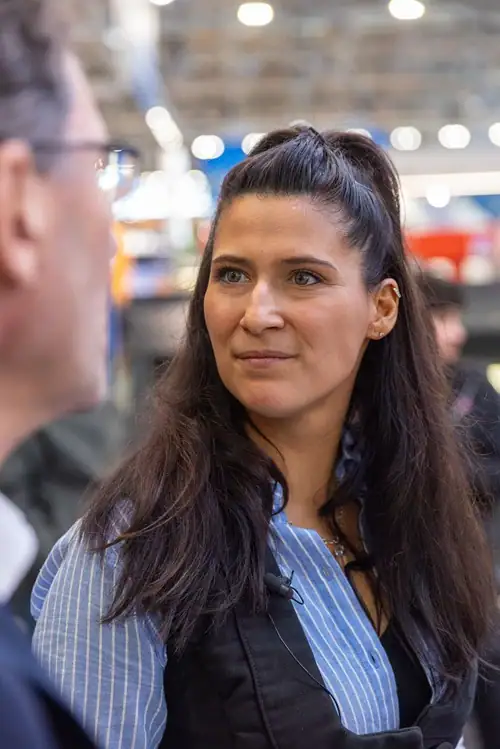 Sonja Theisen mit langen dunklen Haaren im Gespräch mit einem Mann, unscharfer Hintergrund mit Lichtern, drinnen aufgenommen auf der Messe Dach+Holz 2026 in Köln