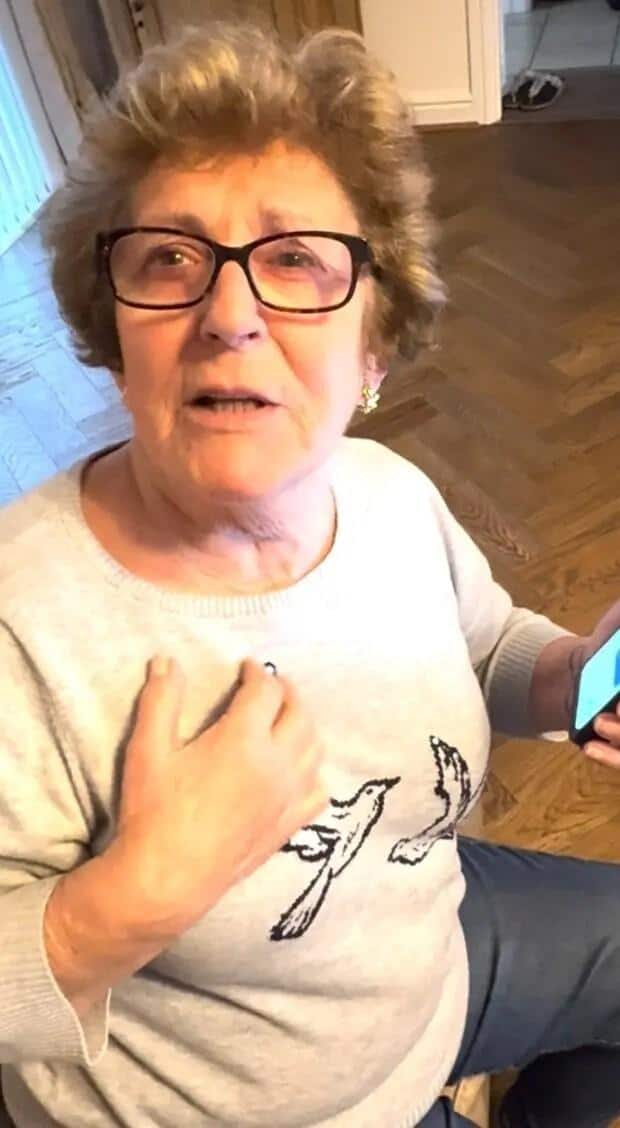 Elderly woman with short, wavy brown hair and glasses, looking concerned. She wears a light gray sweater with bird designs and clutches her chest, likely reacting to a Robert De Niro scam or Facebook fraud. The scene suggests a moment from a "grandma story" about internet deception.