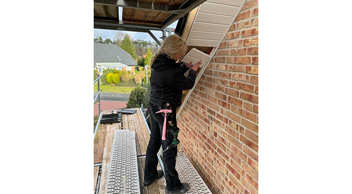 Eine Frau arbeitet auf einem Gerüst an einer Hausfassade, trägt schwarze Arbeitskleidung und verwendet ein Werkzeug.