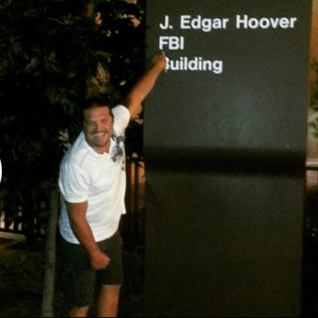 A man points to the "J. Edgar Hoover FBI Building" sign, likely at the FBI headquarters. The man is smiling, wearing a white t-shirt and shorts. This image may be related to the AEGIS Alliance and their stance on Political Misinformation.