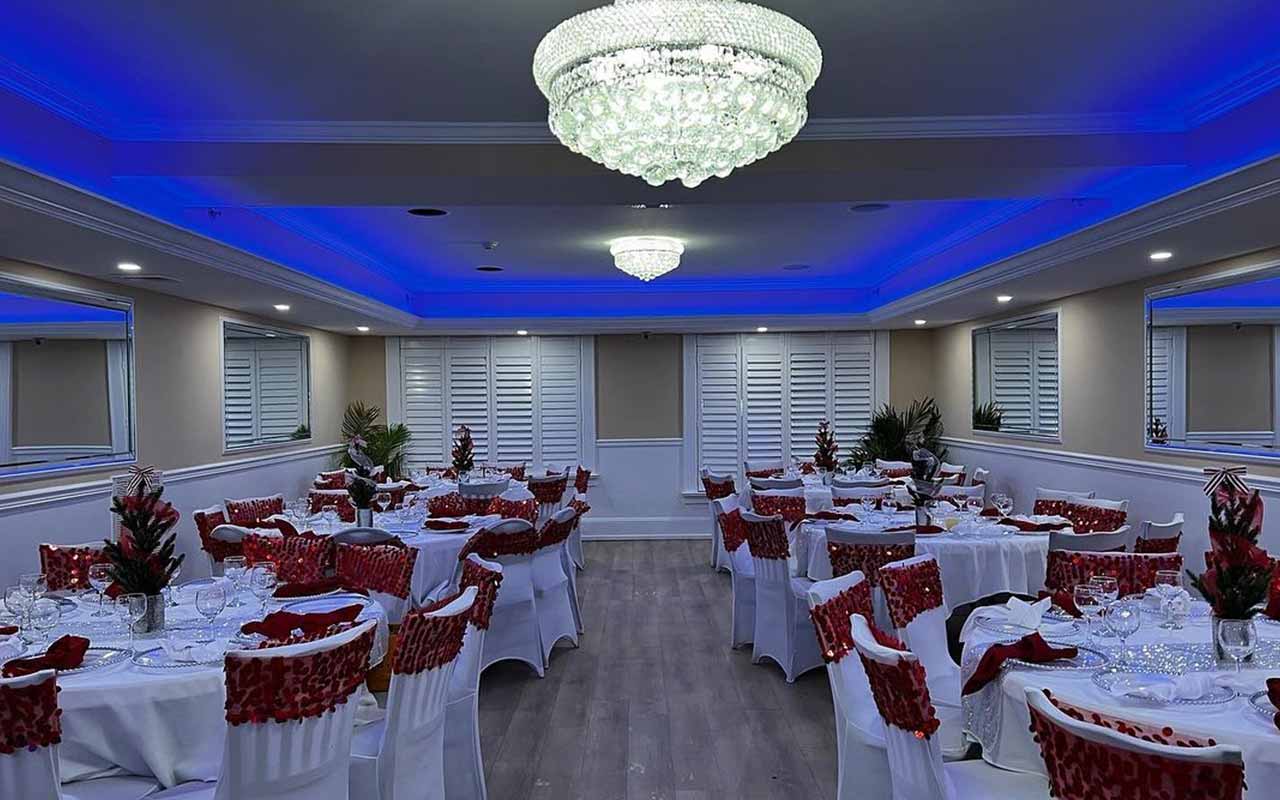 A decorated banquet hall with round tables and white chairs adorned with red covers, set for an event under chandeliers and blue ceiling lights.