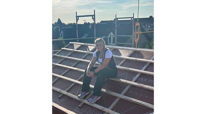 Eine Frau sitzt auf einem Dachgerüst bei sonnigem Wetter und trägt Arbeitskleidung mit Turnschuhen. Im Hintergrund sind weitere Dächer und ein blauer Himmel sichtbar.