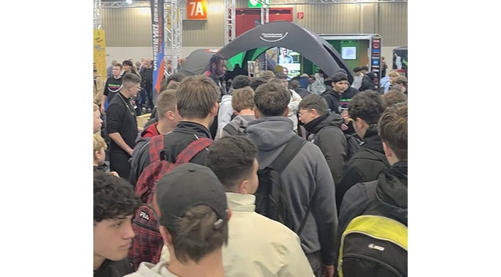 Mehrere Personen stehen dicht gedrängt in einer Schlange auf einer Messe oder Veranstaltung, im Hintergrund sind Stände und ein Zelt sichtbar.