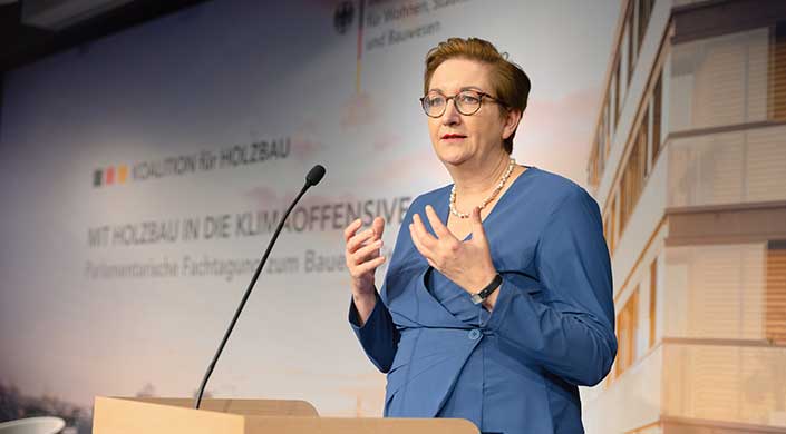 Frau in blauem Anzug und Brille spricht an einem Rednerpult, Hintergrund mit unscharfem Text und deutschem Regierungsemblem.