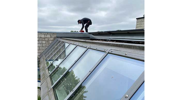 Eine Person arbeitet auf einem Dach bei bewölktem Himmel, neben großen Glasfenstern.