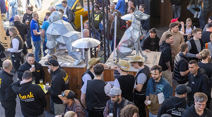 Menschenmenge auf einem belebten Markt; einige tragen Strohhüte und Westen, andere unterhalten sich oder bestellen an einem Essensstand.
