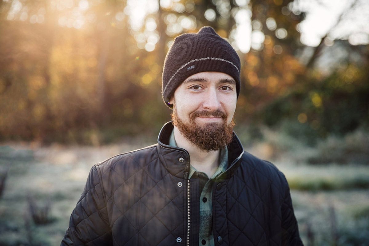 A bearded person wearing a black beanie and quilted jacket stands outdoors with a blurry sunlit background.