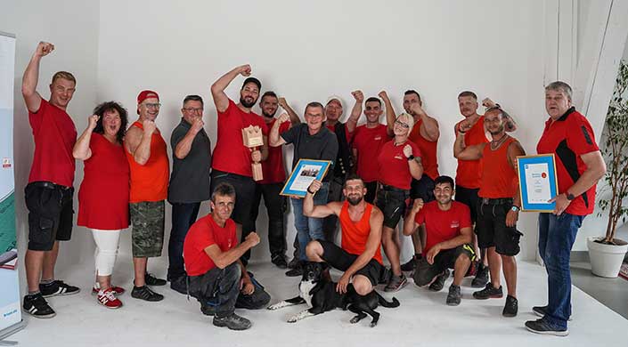 Eine Gruppe von Menschen posiert lächelnd, einige heben die Fäuste, einer hält eine Urkunde und ein Hund sitzt vorne.