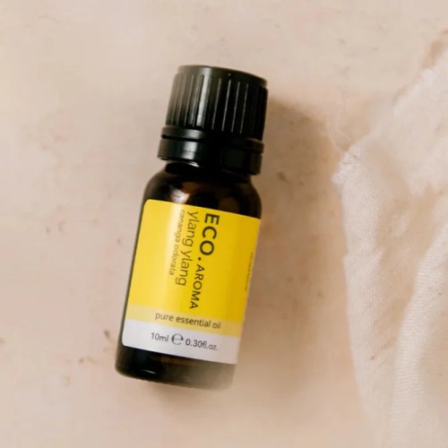 A small dark amber bottle labeled Ylang Ylang Pure Essential Oil sits on a light surface beside a piece of white fabric.