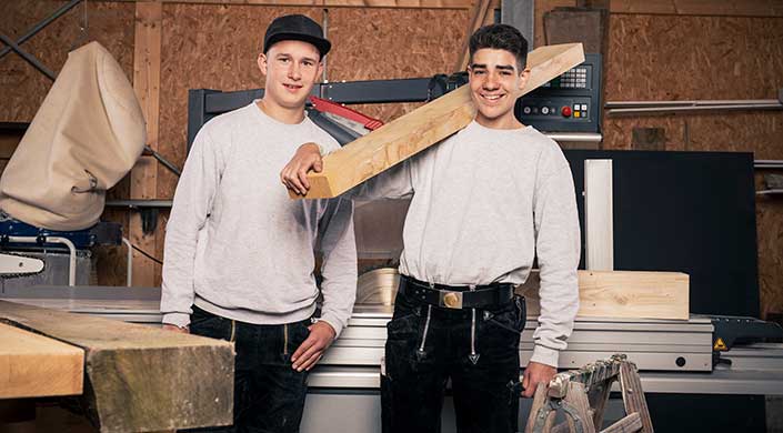 Zwei Männer in Arbeitskleidung stehen in einer Werkstatt, einer hält ein großes Holzbrett vor sich.