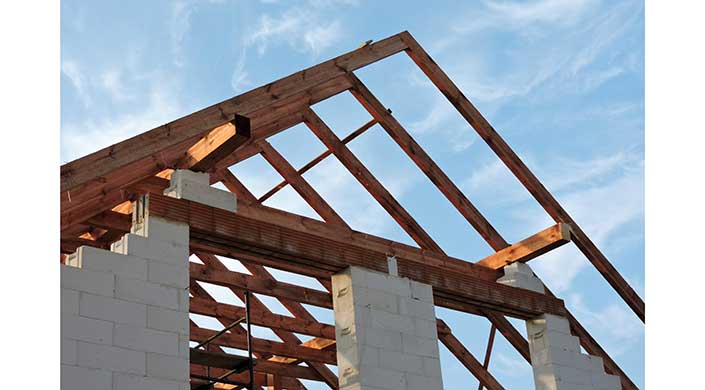 Holzträger bilden das Dachgerüst eines im Bau befindlichen Gebäudes mit weißen Betonblöcken, vor einem blauen Himmel.