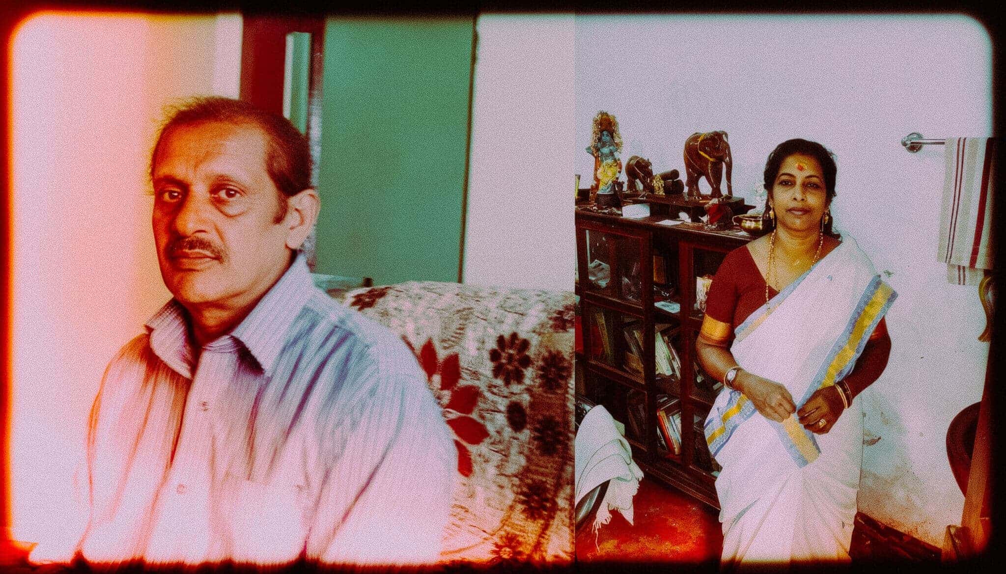 A man seated indoors and a woman in a white sari standing beside a wooden cabinet in a home.