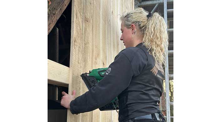 Eine Person benutzt ein grünes Elektrowerkzeug, vermutlich eine Nagelpistole, um Holzplanken an einer vertikalen Holzoberfläche zu befestigen.