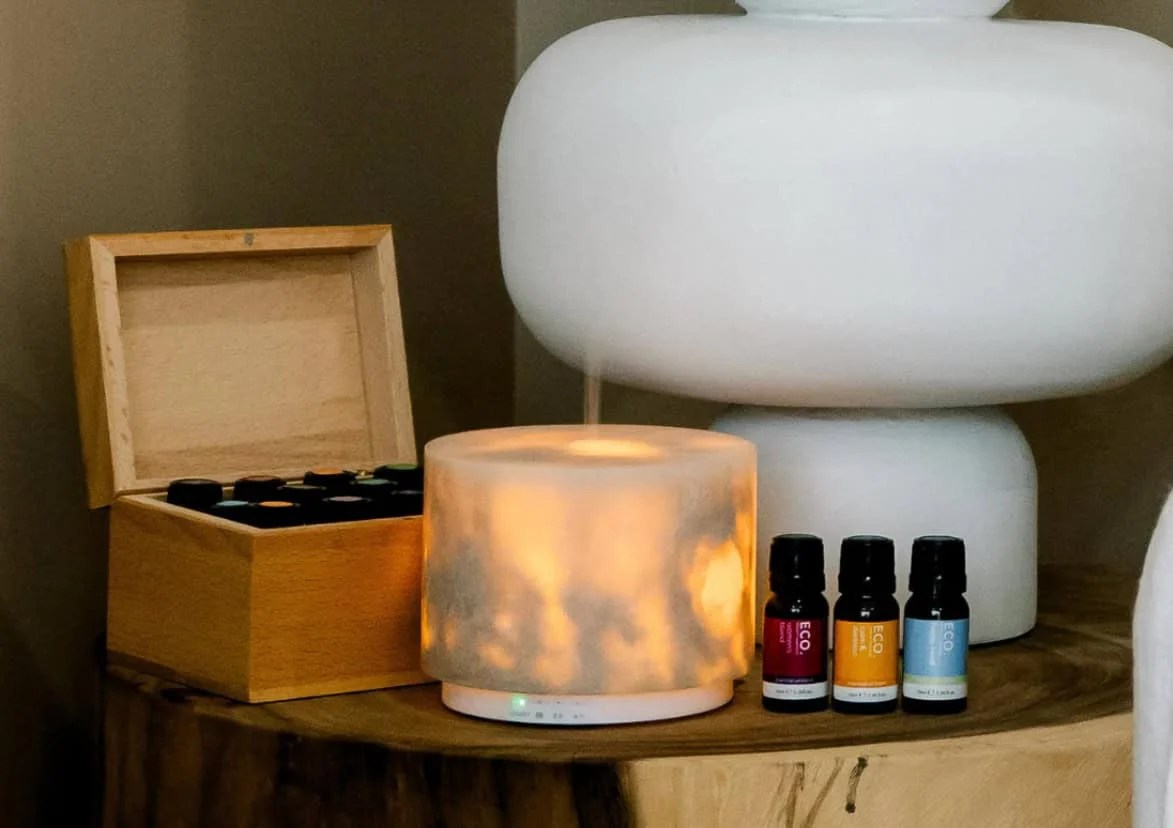 Revive & Thrive Aromatherapy Bundle 10 A lit Marble Diffuser from the Rest & Glow Aromatherapy Bundle emits mist on a wooden surface, accompanied by three essential oils and a large white lamp to enhance the aromatic ambiance.