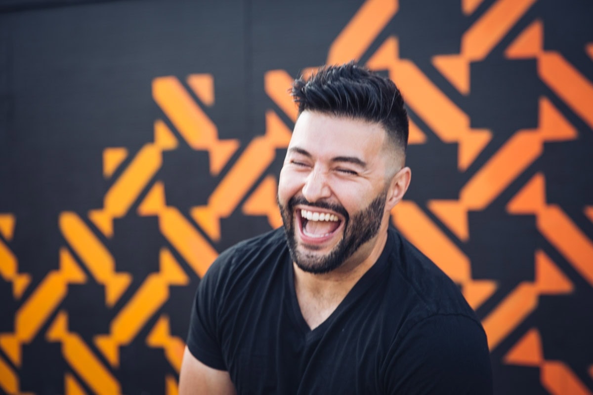 Man with dark hair and beard wearing a black t-shirt is laughing in front of a wall with an orange geometric pattern.