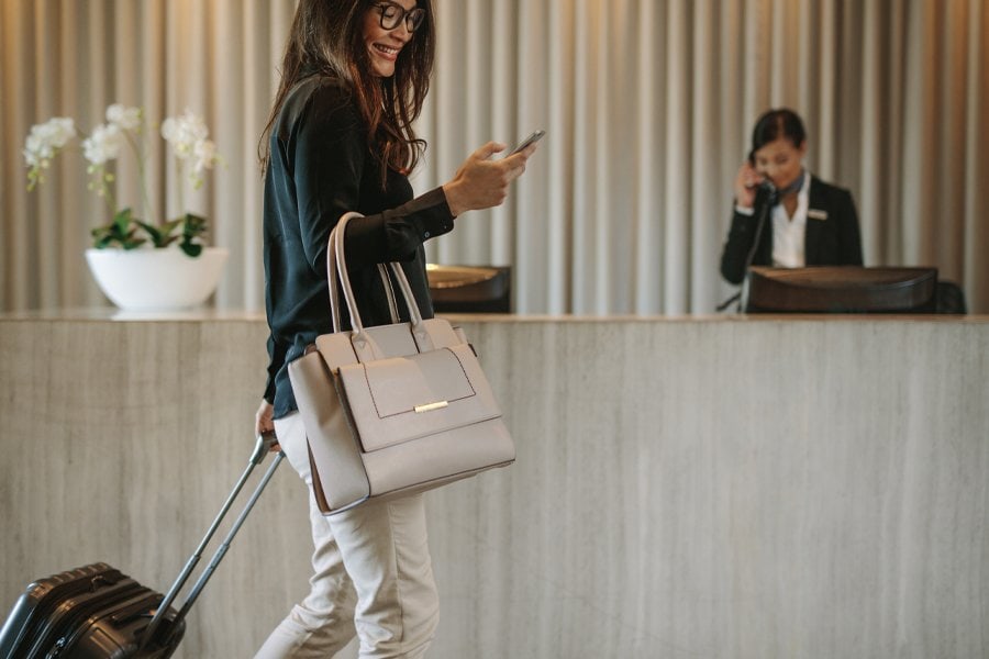 A businesswoman smiling as she walks through a hotel lobby. She is carrying a stylish beige handbag, pulling a compact black suitcase, and using her smartphone. The reception desk is visible in the background, with a hotel staff member on the phone. Orchids in a white vase add a touch of elegance to the minimalist decor.