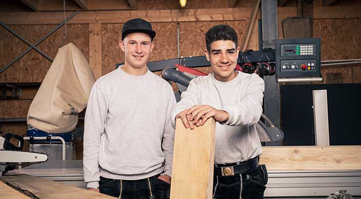 Zwei junge Männer in grauen Pullovern stehen in einer Werkstatt, einer hält ein Holzbrett.
