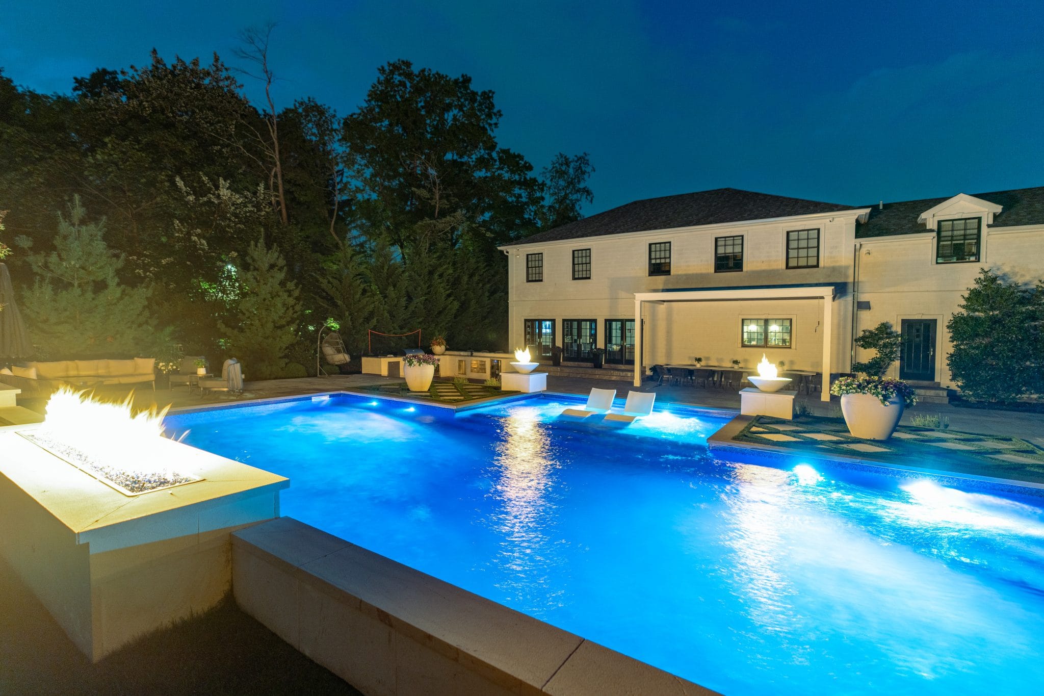 Illuminated backyard pool at night with lounge chairs in the water, fire feature in the foreground, and a large white house in the background.