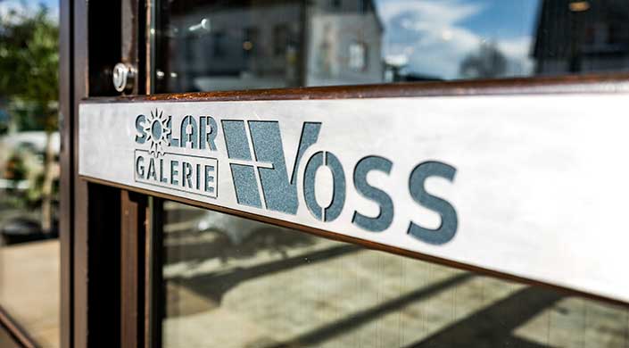 Ein Schild mit dem Text „VOS“ im Vordergrund, unscharfer Hintergrund mit Fenstern und weiteren Details.