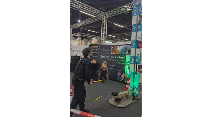 Mann mit Axt steht vor einem Messestand mit Werbebanner, Bau- und Technikthema, moderne Hallenkonstruktion und verschiedene Ausstellungsstücke im Hintergrund.