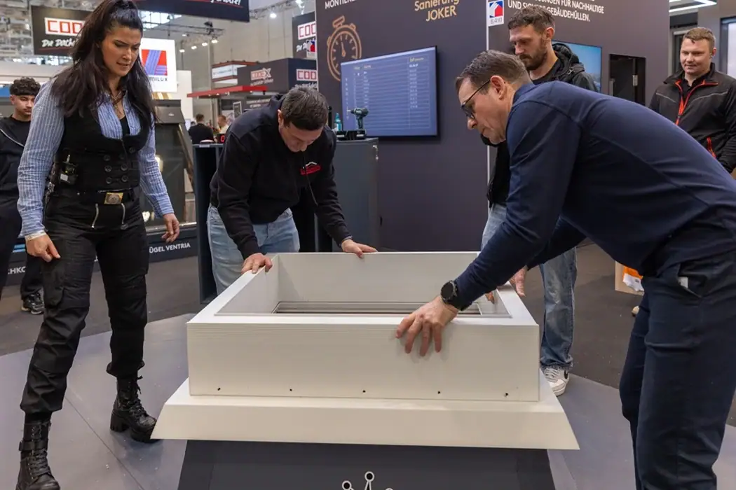 Sonja Theisen, links, coacht zwei Männer, die eine Flachdach-Lichtkuppel so schnell wie möglich aufsetzen und zusammenschrauben, während mehrere Personen zuschauen auf der Messe Dach+Holz 2026 in Köln.