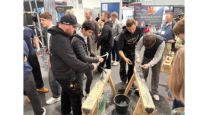 Mehrere Menschen bei einer Veranstaltung; im Vordergrund arbeitet eine Person mit Werkzeug an einem Werkstück, während andere im Hintergrund stehen und sich unterhalten.