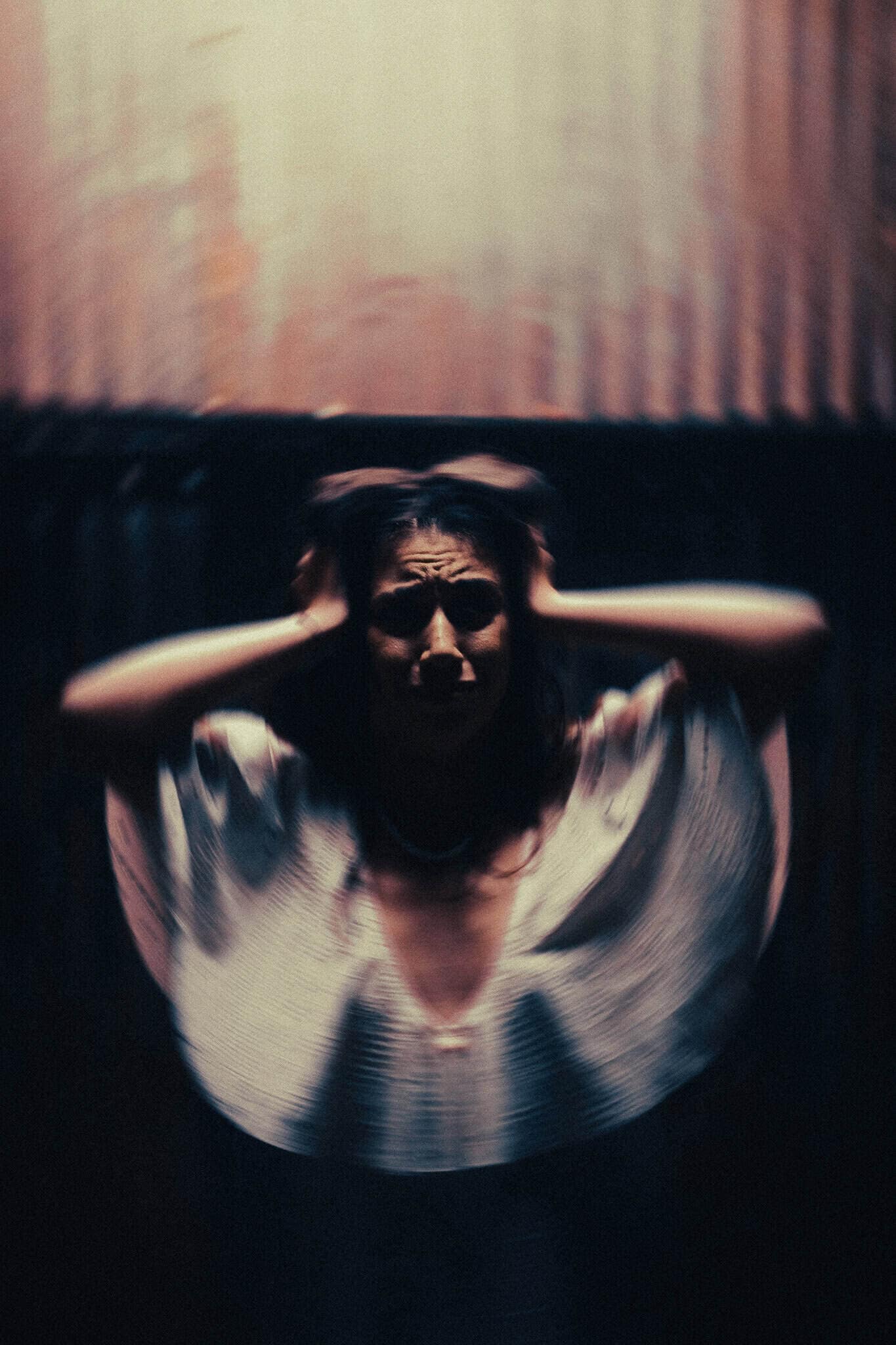 A blurred figure sits in a dim room holding both hands against the sides of their head.