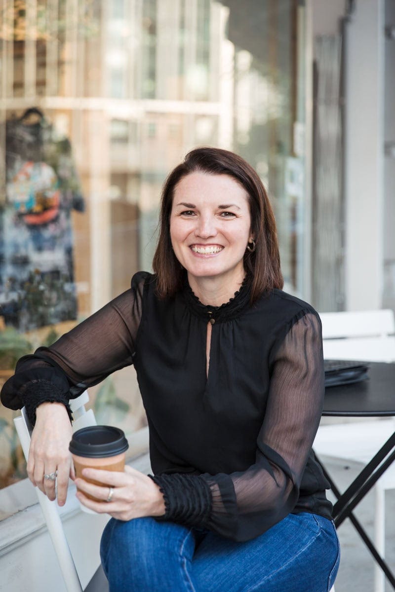 Woman sitting outdoors with a coffee cup, wearing a black blouse and jeans, smiling at the camera.