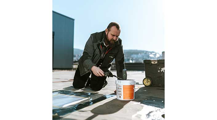 Ein Mann kniet auf dem Boden und arbeitet mit Werkzeug und einem Eimer auf einer Baustelle im Freien.