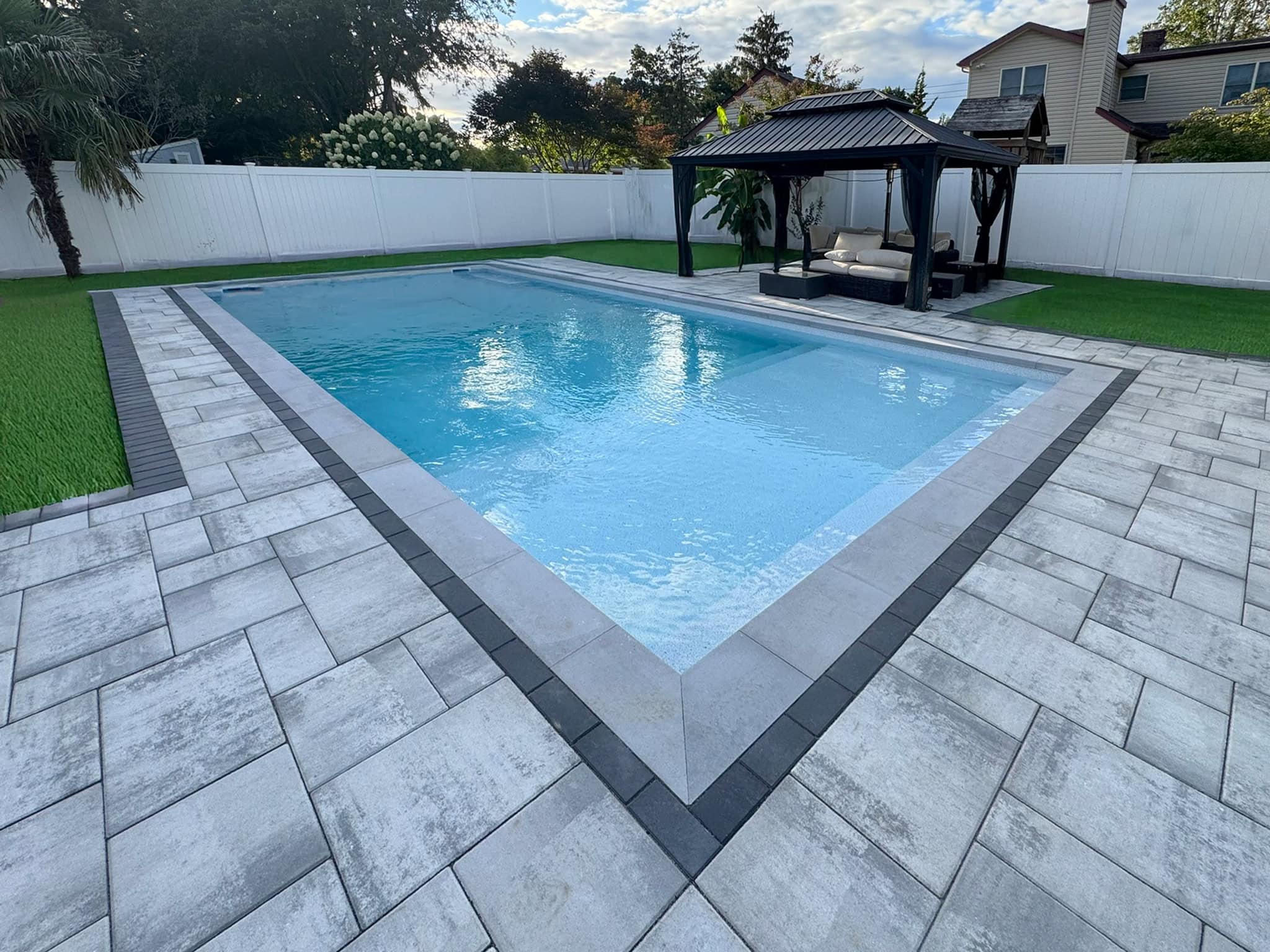 Rectangular outdoor swimming pool with light gray tile deck, surrounded by artificial grass and a white fence, with a black gazebo and lounge chairs in the background.