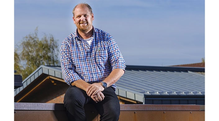 Ein Mann mit kariertem Hemd sitzt draußen, teilweise abgeschnitten, Hintergrund mit blauem Himmel und modernem Gebäude.
