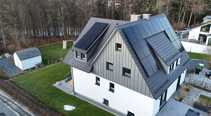 Modernes Haus mit grauem Dach und Solarpaneelen, weißen und grauen Fassadenelementen, großen Fenstern und grünem Rasen im Vordergrund, Wald im Hintergrund.