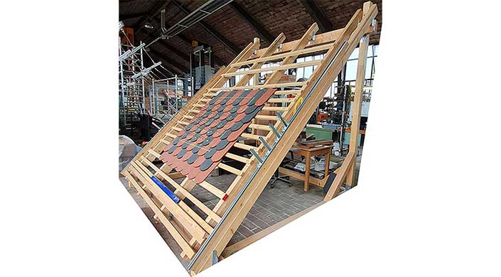 Bild von Windsog im Institut für Ziegelforschung Holzgerüst mit geneigtem Dach, das halbfertig mit roten und schwarzen Dachziegeln gedeckt ist, in einer Werkstattumgebung.