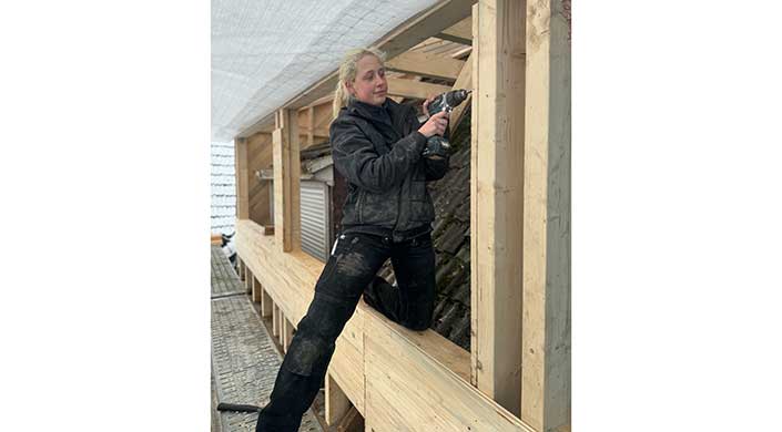 Eine Person mit blonden Haaren arbeitet auf einer Baustelle und benutzt eine elektrische Bohrmaschine, trägt Arbeitskleidung und ist von Holzgerüsten umgeben.
