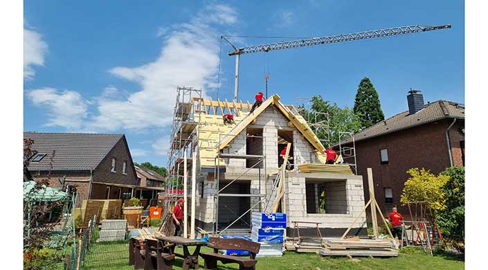 Arbeiter bauen ein Hausdach, Gerüst steht am Rohbau, Baukran im Hintergrund, blauer Himmel, Baumaterialien und Holzlatten sichtbar.