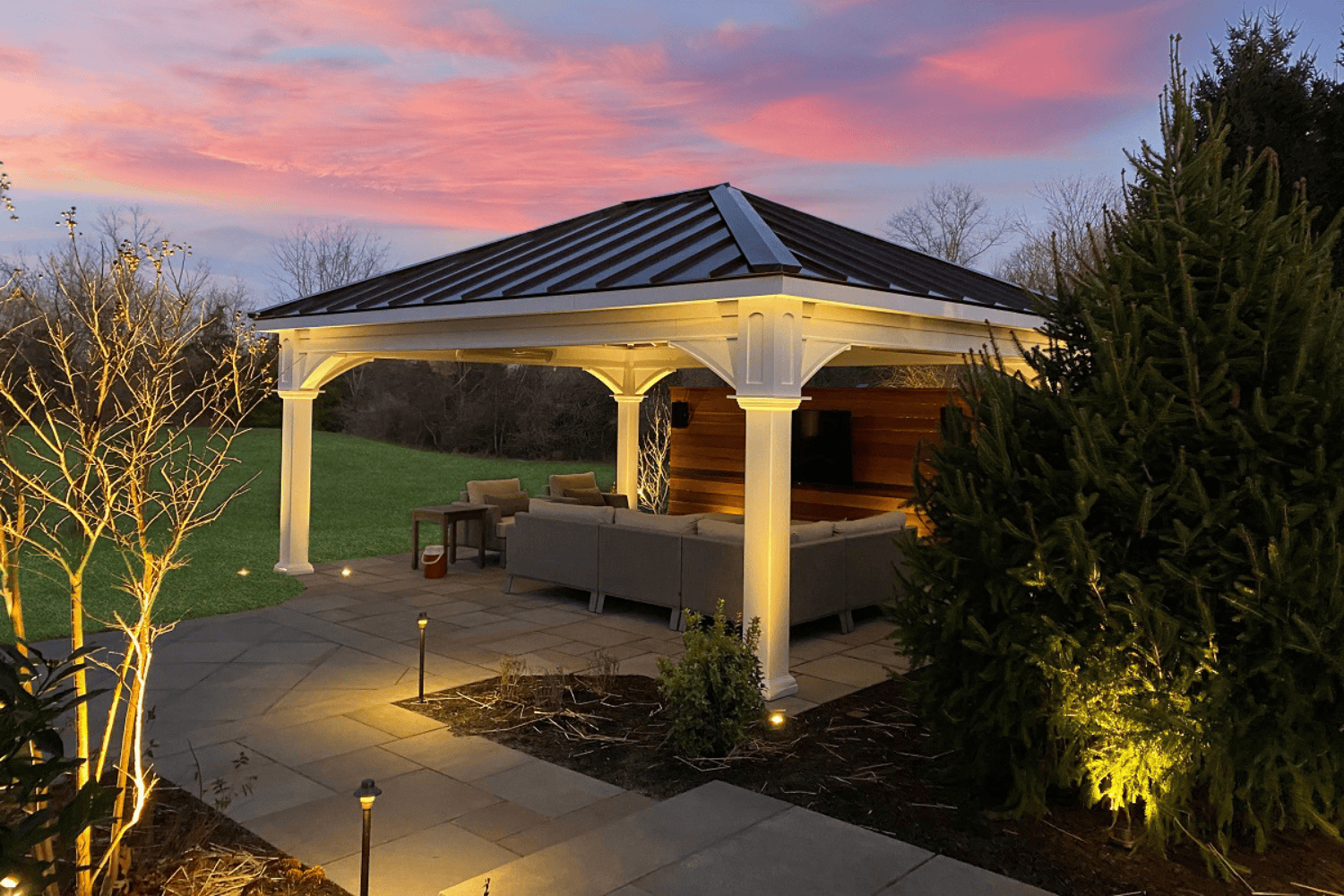 A modern pavilion with outdoor seating is illuminated at dusk, set on a stone patio with surrounding trees and landscaping under a colorful sky.