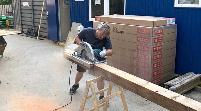 Eine Person benutzt eine Kreissäge, um ein Holzbrett zu schneiden, während sie auf einer Baustelle arbeitet. Im Hintergrund sind Kartons zu sehen.
