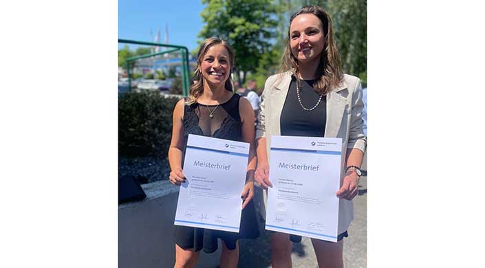 Zwei Frauen stehen draußen und halten Meisterbriefe, lächeln in die Kamera, sommerliches Wetter mit grünen Bäumen im Hintergrund.