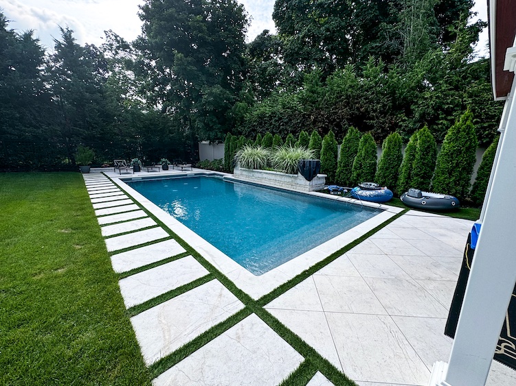 Rectangular outdoor swimming pool with white stone paving, green lawn, and hedges surrounding the area; pool floats are on the poolside.